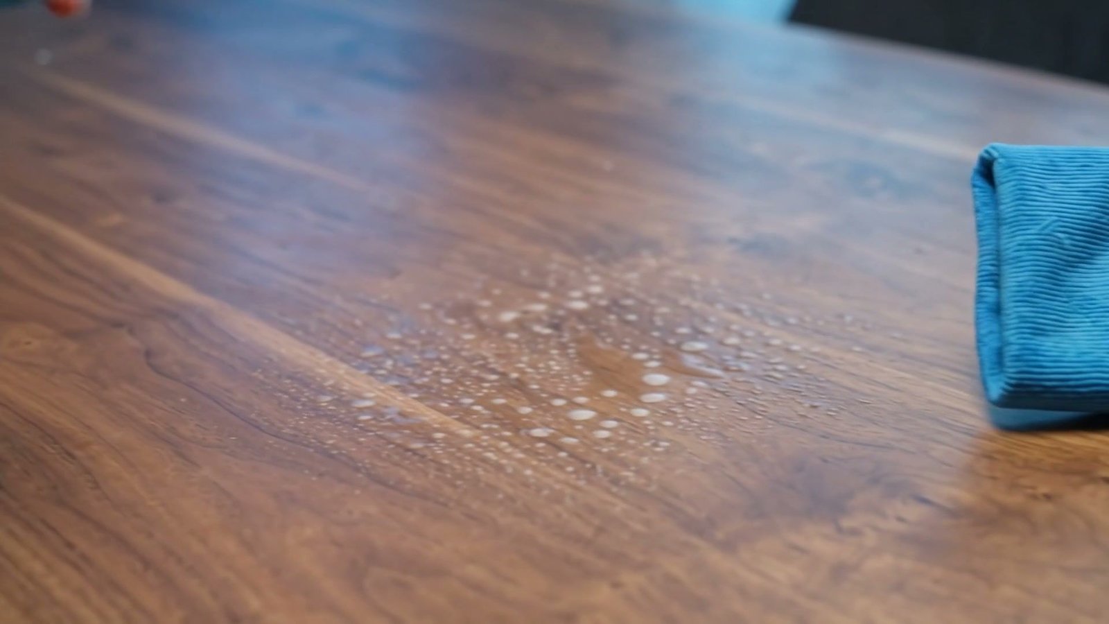 A blue microfiber cloth wiping cleaning spray off a wooden table